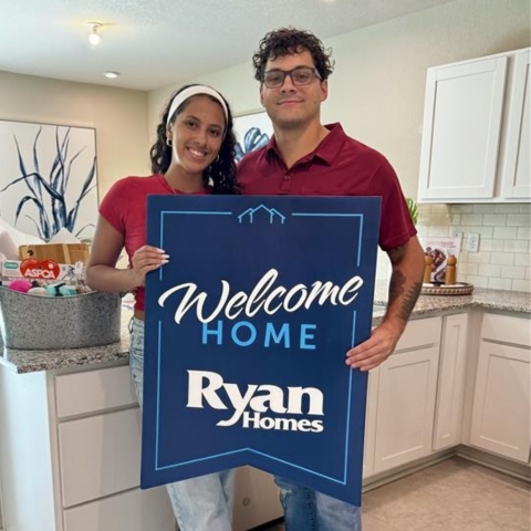 Couple celebrating move-in day in their new Ryan Homes kitchen, holding a 'Welcome Home Ryan Homes' sign